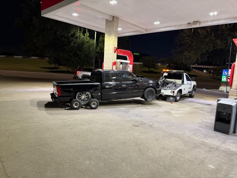 A black lowered truck being towed at a gas station by a white tow truck during the night.