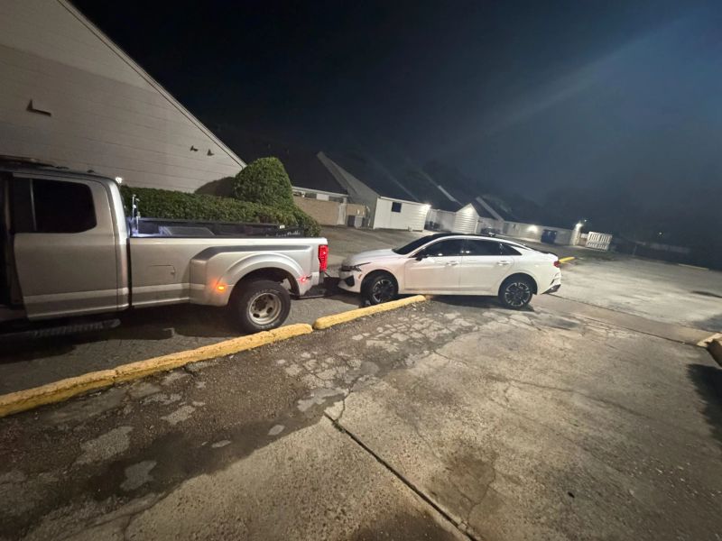 A white car parked awkwardly on a curb beside a silver pickup truck, indicating a towing situation at night.