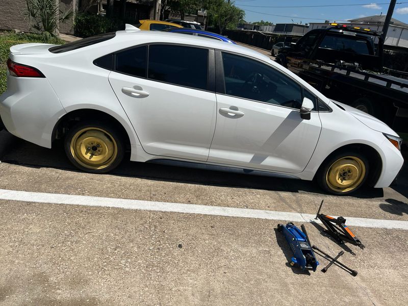 White car with a flat tire and yellow spare wheel being towed, accompanied by jacks.
