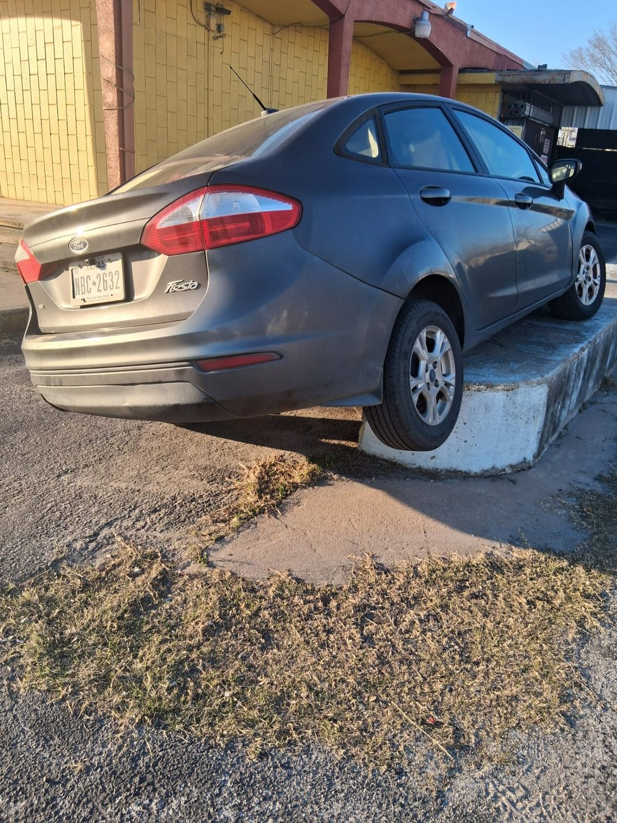 A grey Ford Fiesta stuck awkwardly on a concrete curb in Houston, indicating a possible need for winch out, towing or assistance.
