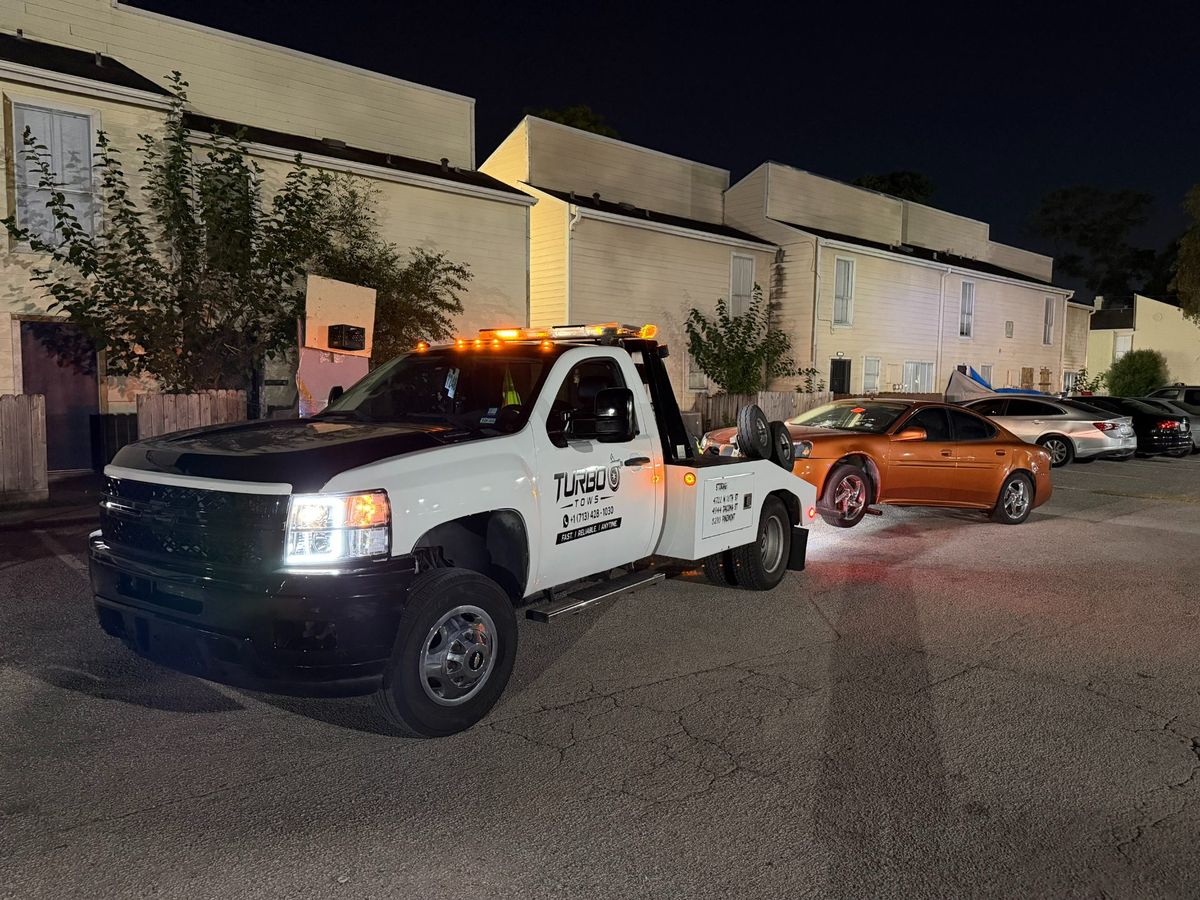 Turbo Tows tow truck assisting an orange car in a parking lot at night in Houston.