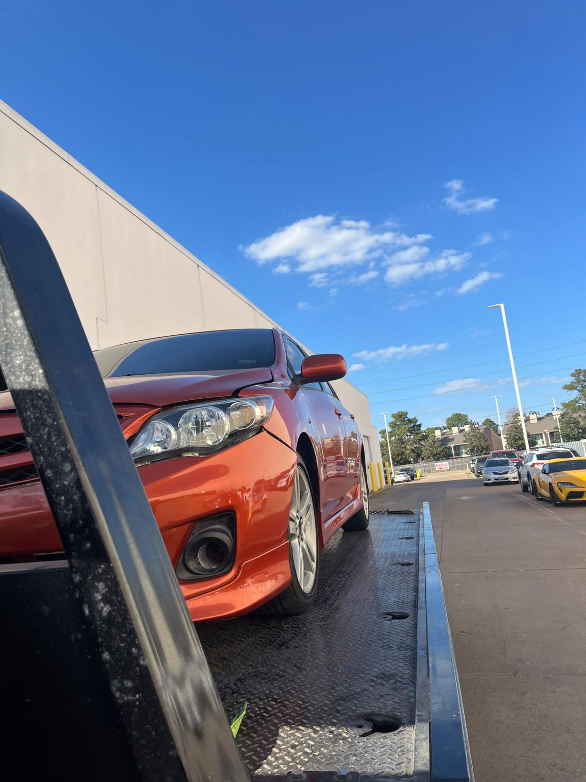 Orange car being towed on a flatbed truck in Houston, Texas under a clear blue sky.