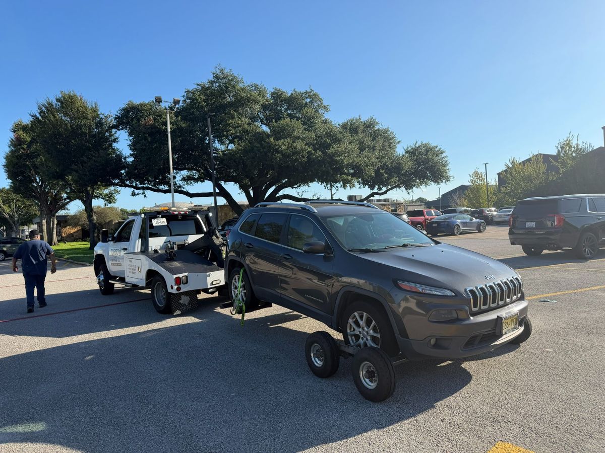 A gray SUV being towed by a truck in a parking lot, with clear blue skies and trees in the background.